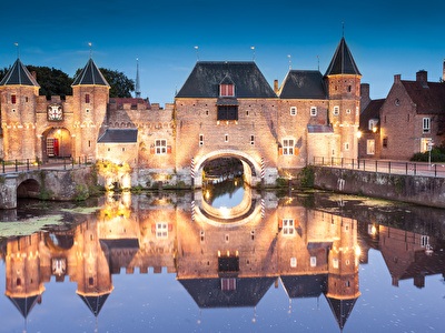 Een kasteel met een brug over water, omringd door bomen, die zich reflecteren in het water, met de prachtig verlichte structuur en de weerspiegeling in de gracht of het meer.