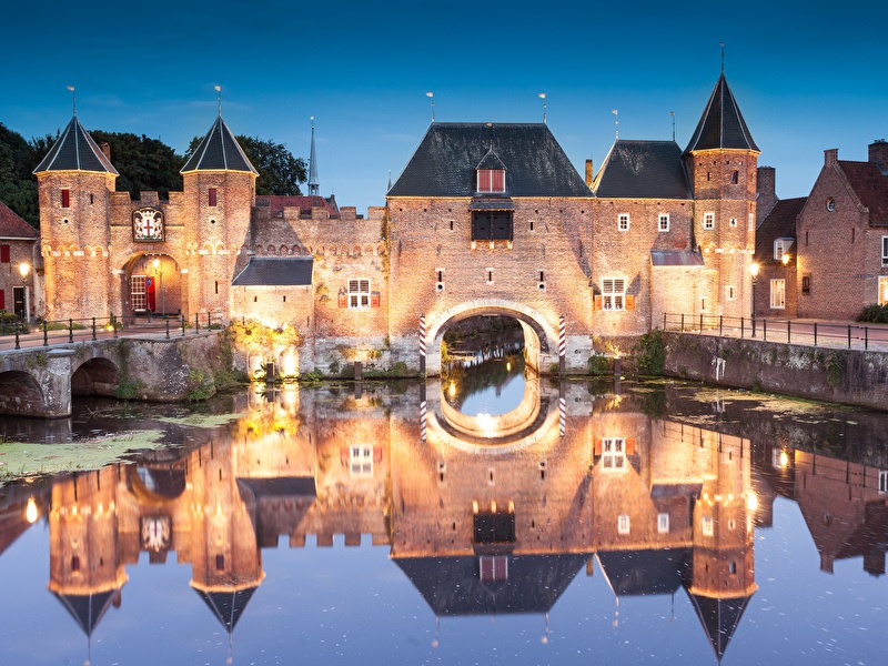 Een kasteel met een brug over water, omringd door bomen, die zich reflecteren in het water, met de prachtig verlichte structuur en de weerspiegeling in de gracht of het meer.